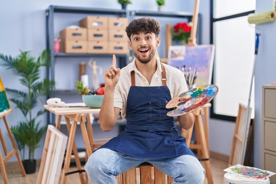 Arab man with beard painter sitting at art studio holding palette surprised with an idea or question pointing finger with happy face, number one