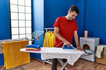 Young hispanic man smiling confident ironing clothes at laundry room