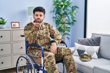 Fototapeta premium Arab man wearing camouflage army uniform sitting on wheelchair looking at the camera blowing a kiss with hand on air being lovely and sexy. love expression.