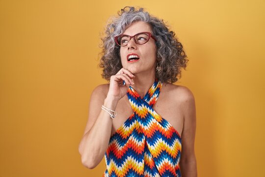 Middle age woman with grey hair standing over yellow background with hand on chin thinking about question, pensive expression. smiling with thoughtful face. doubt concept.