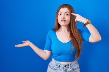 Redhead woman standing over blue background confused and annoyed with open palm showing copy space and pointing finger to forehead. think about it.