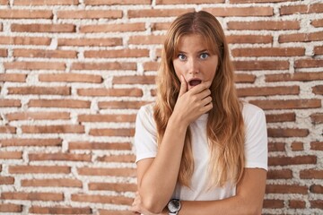 Fototapeta na wymiar Young caucasian woman standing over bricks wall looking fascinated with disbelief, surprise and amazed expression with hands on chin