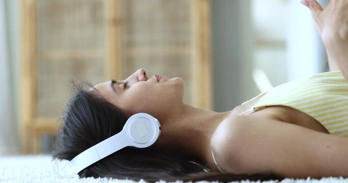 Peaceful engaged pretty young girl in big wireless headphones resting on soft carpeted floor, listening to music, singing song, enjoying leisure, rhythm, relaxation