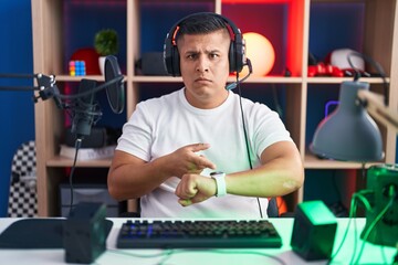 Young hispanic man playing video games in hurry pointing to watch time, impatience, upset and angry...