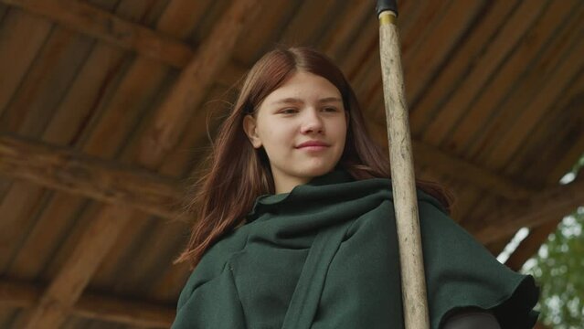 Smiling girl in dark green cloak stands in gazebo. Young female warrior holds spear and waits for fighting training on blurred background close low angle shot
