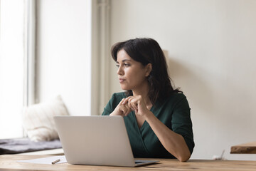 Serious woman sit at workplace desk with laptop starting aside, search solution stuck with difficult task, faced with business challenge, thinks over task, lack of understanding, motivation or ideas