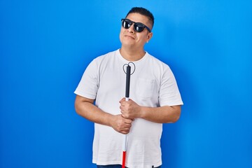 Hispanic young blind man holding cane smiling looking to the side and staring away thinking.