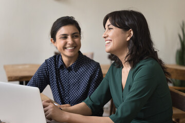 Indian and Armenian women talking, working together on project having friendly conversation, share ideas engaged in creative team-work use laptop seated at desk, communication, teamwork, friendship