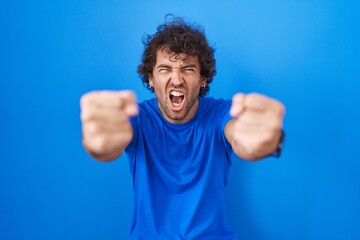 Fototapeta premium Hispanic young man standing over blue background angry and mad raising fists frustrated and furious while shouting with anger. rage and aggressive concept.