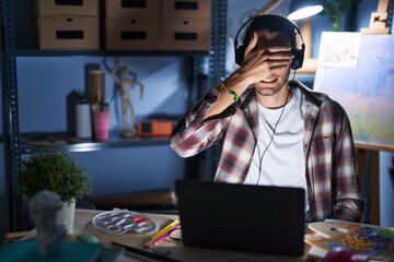 Young hispanic man sitting at art studio with laptop late at night smiling and laughing with hand on face covering eyes for surprise. blind concept.