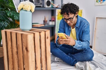 Young hispanic man artist smiling confident using smartphone at art studio