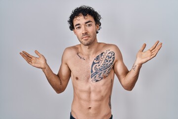 Hispanic man standing shirtless clueless and confused expression with arms and hands raised. doubt...