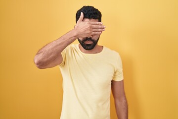 Fototapeta na wymiar Hispanic man with beard standing over yellow background covering eyes with hand, looking serious and sad. sightless, hiding and rejection concept