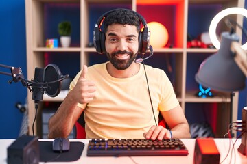 Hispanic man with beard playing video games with headphones doing happy thumbs up gesture with...