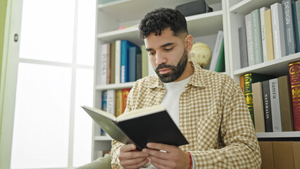 Fototapeta premium Young hispanic man student reading book at library university