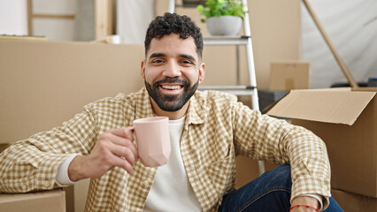 Young hispanic man drinking coffee sitting on floor at new home