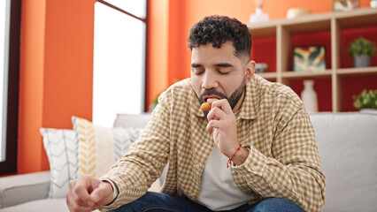 Young hispanic man eating doughnut sitting on sofa at h