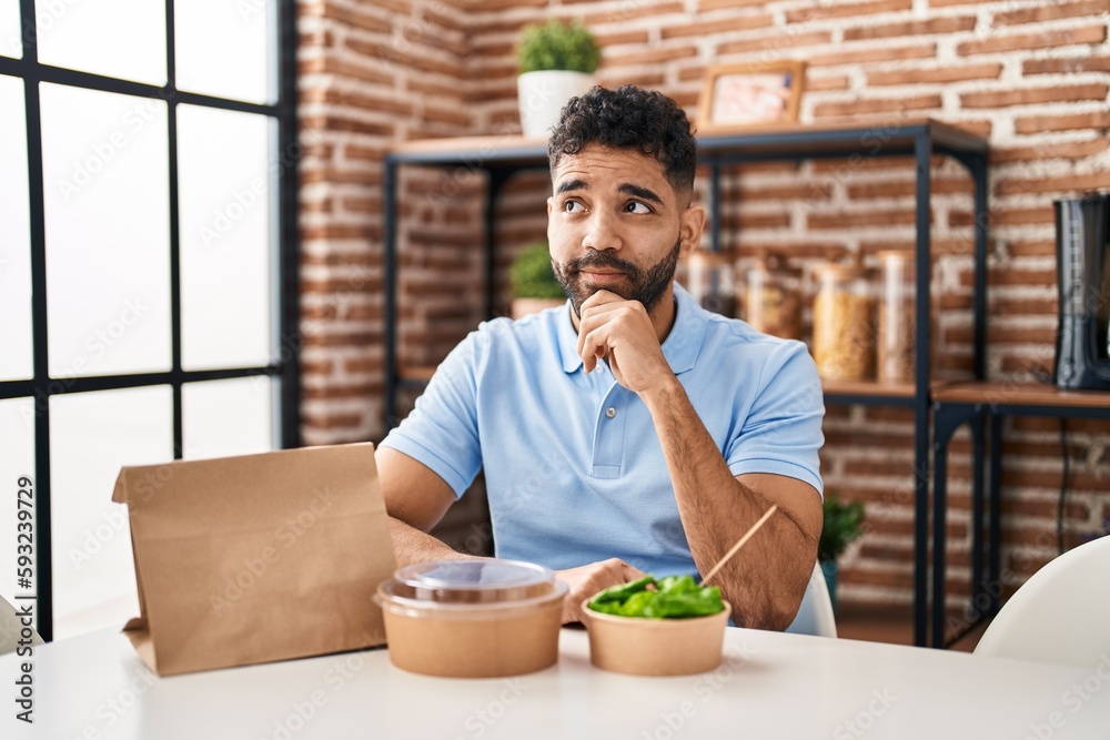 Wall mural Hispanic man with beard eating delivery salad serious face thinking about question with hand on chin, thoughtful about confusing idea