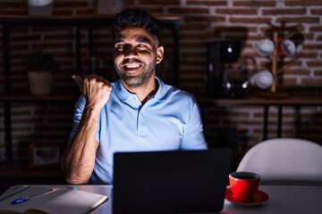 Hispanic man with beard using laptop at night smiling with happy face looking and pointing to the side with thumb up.