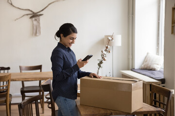 Millennial Indian woman sending cardboard box, prepare goods for shipment using smartphone, calling...