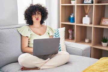 Young brunette woman with curly hair using laptop sitting on the sofa at home puffing cheeks with funny face. mouth inflated with air, crazy expression.