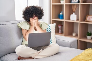 Young brunette woman with curly hair using laptop sitting on the sofa at home with sad expression covering face with hands while crying. depression concept.