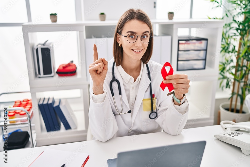 Sticker Young caucasian doctor woman holding support red ribbon surprised with an idea or question pointing finger with happy face, number one