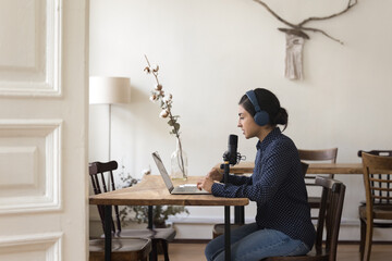 Focused young Indian woman in wireless headphones look at laptop screen, talking in professional microphone on stand, record vlog or new podcast, share experience with internet audience at home office