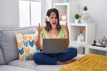 Hispanic young woman using laptop at home shouting with crazy expression doing rock symbol with hands up. music star. heavy concept.