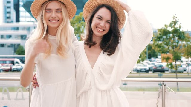 Two young beautiful smiling hipster female in trendy summer white dress clothes and straw hats. Sexy carefree women walking in street. Positive models having fun, hugging and laughing 