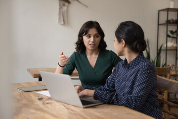 Two young office workers, female students sit at desk with laptop, share ideas, talk, discuss...