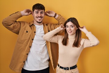Young hispanic couple standing over yellow background smiling pulling ears with fingers, funny...