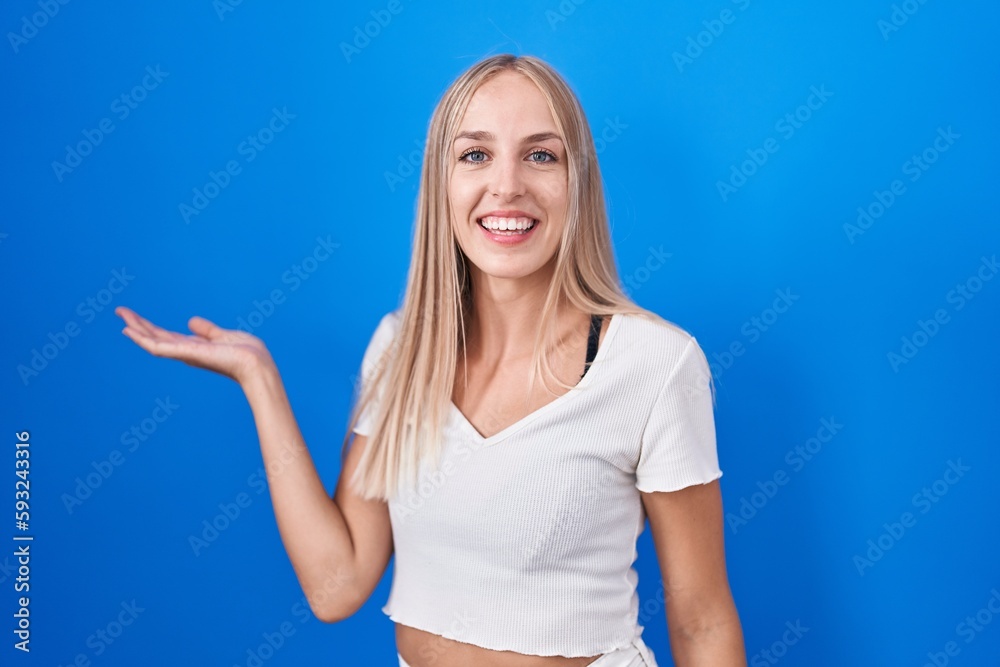 Poster young caucasian woman standing over blue background smiling cheerful presenting and pointing with pa