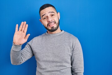 Hispanic man standing over blue background waiving saying hello happy and smiling, friendly welcome gesture © Krakenimages.com