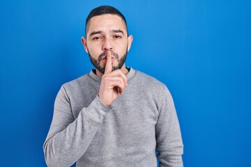 Hispanic man standing over blue background asking to be quiet with finger on lips. silence and secret concept.