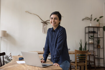 Portrait of confident Indian female employee, freelancer, student smile look at cam, standing at workplace with laptop, enjoy professional achievement, career growth or promotion, use new app for work