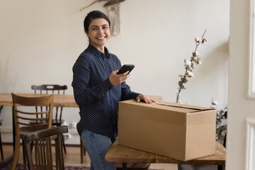 Satisfied Indian woman holds smartphone prepare postal parcel box for sending enjoy express...