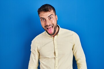 Handsome hispanic man standing over blue background sticking tongue out happy with funny expression. emotion concept.