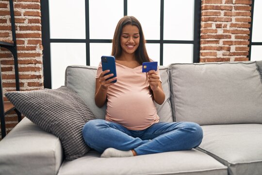 Young latin woman pregnant using smartphone and credit card at home