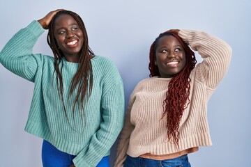 Two african woman standing over blue background smiling confident touching hair with hand up gesture, posing attractive and fashionable