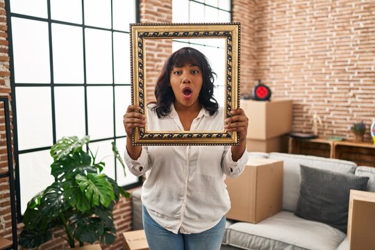 Hispanic woman at new home holding empty frame afraid and shocked with surprise and amazed expression, fear and excited face.
