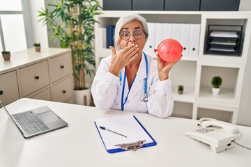 Middle age woman with grey hair wearing doctor uniform holding balloon covering mouth with hand, shocked and afraid for mistake. surprised expression