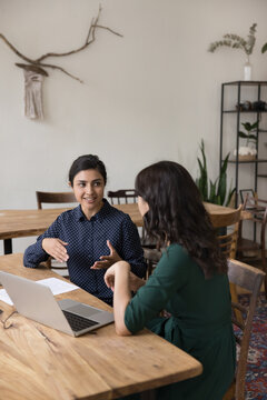 Millennial Indian woman explains to colleague details of collaborative project sit together at desk share ideas, talking, making joint task. Sales manager meet with client, offering company services