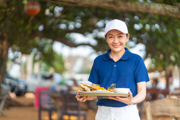 Portrait of Asian woman waiter serving food and drink to customer on the table at tropical beach cafe and restaurant on summer holiday vacation. Food and drink business service occupation concept.