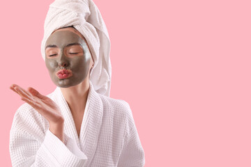 Young woman with applied clay mask blowing kiss on pink background