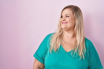 Caucasian plus size woman standing over pink background looking away to side with smile on face, natural expression. laughing confident.