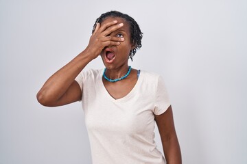 African woman with dreadlocks standing over white background peeking in shock covering face and eyes with hand, looking through fingers with embarrassed expression.