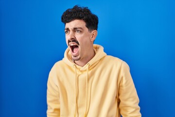 Fototapeta na wymiar Hispanic man standing over blue background angry and mad screaming frustrated and furious, shouting with anger. rage and aggressive concept.