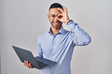 Young hispanic man working using computer laptop doing ok gesture with hand smiling, eye looking through fingers with happy face.