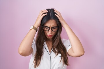 Young brunette woman wearing glasses standing over pink background suffering from headache desperate and stressed because pain and migraine. hands on head.